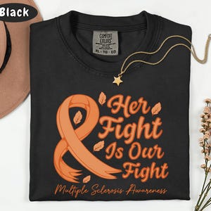 May include: Black t-shirt with an orange ribbon and the text "Her Fight Is Our Fight" in orange script. The shirt also includes the text "Multiple Sclerosis Awareness". A gold necklace with a star pendant is visible.