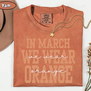 May include: A burnt orange t-shirt with the text "IN MARCH WE WEAR ORANGE" in a layered design. A gold necklace with a star pendant rests on the shirt. A brown hat is visible in the upper left corner.