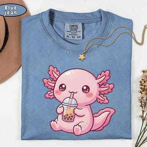 May include: Blue jean Comfort Colors t-shirt featuring a pink axolotl cartoon drinking boba tea. The axolotl has large, expressive eyes and pink frills. A gold necklace with a star pendant is on the shirt. The shirt is size XL.