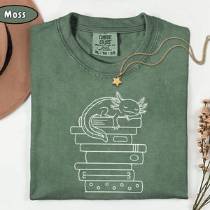 May include: A moss green t-shirt with a white axolotl graphic sleeping on a stack of books. The shirt has a crew neck and short sleeves. A gold necklace with a star pendant is draped on the shirt.