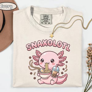 以下が含まれることがあります： アイボリー色のTシャツ。漫画のウーパールーパーのデザイン。ピンク色のウーパールーパーがラーメンを食べ、タピオカティーを持っています。「SNAXOLOTL」の文字がウーパールーパーの上にあります。星のペンダントが付いたゴールドのネックレスがシャツの上にあります。