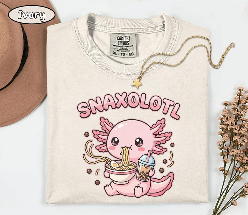 以下が含まれることがあります： アイボリー色のTシャツ。漫画のウーパールーパーのデザイン。ピンク色のウーパールーパーがラーメンを食べ、タピオカティーを持っています。「SNAXOLOTL」の文字がウーパールーパーの上にあります。星のペンダントが付いたゴールドのネックレスがシャツの上にあります。