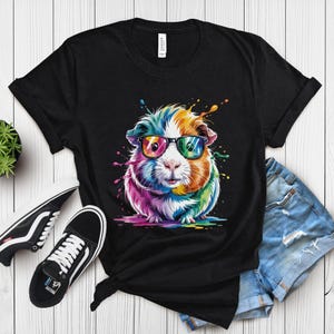 May include: Black t-shirt featuring a colorful guinea pig wearing sunglasses. The guinea pig is depicted with vibrant rainbow hues and paint splatter effects. The shirt is paired with black sneakers and denim shorts, creating a casual, fun look.