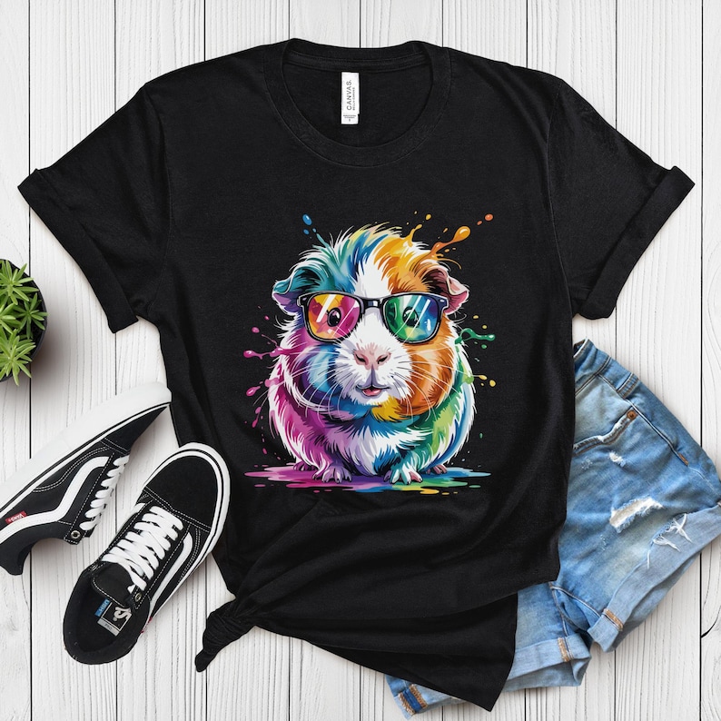 May include: Black t-shirt featuring a colorful guinea pig wearing sunglasses. The guinea pig is depicted with vibrant rainbow hues and paint splatter effects. The shirt is paired with black sneakers and denim shorts, creating a casual, fun look.