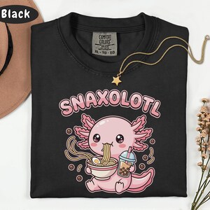以下が含まれることがあります： 黒いTシャツ。ピンクと白のアホロートルがラーメンを食べ、バブルティーを持っているグラフィック。テキスト「Snaxolotl」はアホロートルの上にあります。星のペンダントが付いた金のネックレスがシャツにあります。