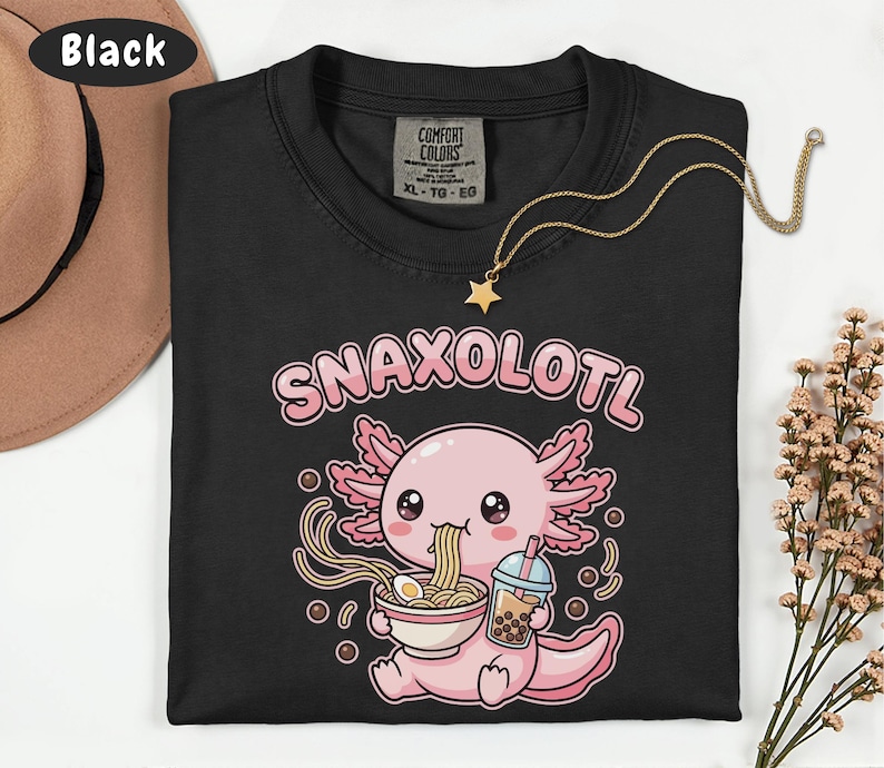 以下が含まれることがあります： 黒いTシャツ。ピンクと白のアホロートルがラーメンを食べ、バブルティーを持っているグラフィック。テキスト「Snaxolotl」はアホロートルの上にあります。星のペンダントが付いた金のネックレスがシャツにあります。