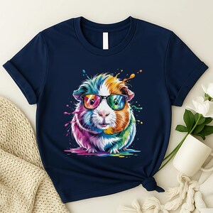 May include: Navy blue t-shirt featuring a colorful guinea pig wearing sunglasses. The guinea pig has a rainbow-colored coat with splashes of paint around it. The t-shirt is knotted at the bottom.