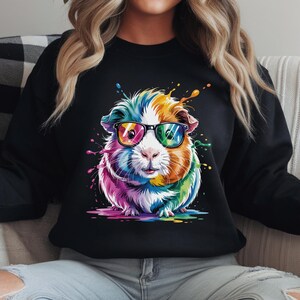 May include: Black sweatshirt featuring a colorful guinea pig wearing sunglasses. The guinea pig is depicted with vibrant rainbow hues and paint splatter effects. The design is centered on the front of the sweatshirt.