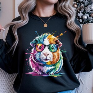 May include: Black sweatshirt featuring a colorful guinea pig wearing glasses. The guinea pig is depicted with rainbow-colored fur and a playful expression. Splashes of paint in various colors surround the guinea pig, adding to the whimsical design.