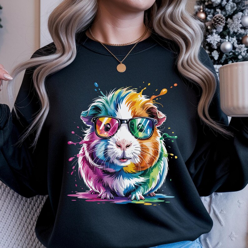 May include: Black sweatshirt featuring a colorful guinea pig wearing glasses. The guinea pig is depicted with rainbow-colored fur and a playful expression. Splashes of paint in various colors surround the guinea pig, adding to the whimsical design.
