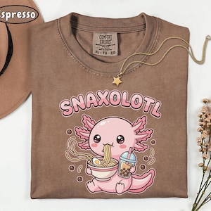 以下が含まれることがあります： 「Snaxolotl」の文字と、ラーメンを食べ、飲み物を持っている漫画のウーパールーパーが描かれた茶色のTシャツ。星のペンダントが付いたゴールドのネックレスがシャツの上にあります。シャツには「Comfort Colors」と書かれており、サイズはXLです。