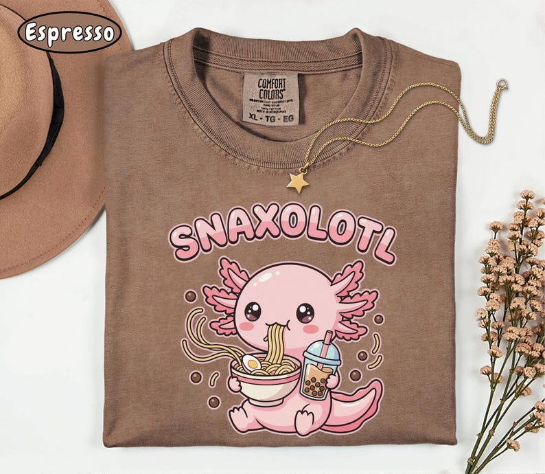 以下が含まれることがあります： 「Snaxolotl」の文字と、ラーメンを食べ、飲み物を持っている漫画のウーパールーパーが描かれた茶色のTシャツ。星のペンダントが付いたゴールドのネックレスがシャツの上にあります。シャツには「Comfort Colors」と書かれており、サイズはXLです。