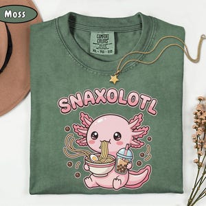以下が含まれることがあります： モスグリーンのTシャツ。ピンク色のウーパールーパーがラーメンを食べ、ボバドリンクを持っているグラフィック。テキスト「SNAXOLOTL」はウーパールーパーの上にあります。星のペンダントが付いたゴールドのネックレスがシャツの上にあります。
