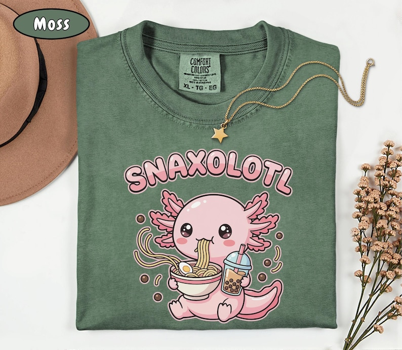 以下が含まれることがあります： モスグリーンのTシャツ。ピンク色のウーパールーパーがラーメンを食べ、ボバドリンクを持っているグラフィック。テキスト「SNAXOLOTL」はウーパールーパーの上にあります。星のペンダントが付いたゴールドのネックレスがシャツの上にあります。