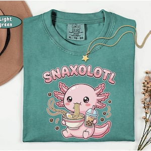 以下が含まれることがあります： 「Snaxolotl」の文字と、ラーメンを食べ、ボバを飲んでいる漫画のウーパールーパーが描かれた、ライトグリーンのComfort Colors Tシャツ。 金色の星のネックレスがシャツの上にかけられています。 茶色の帽子が左上にあります。