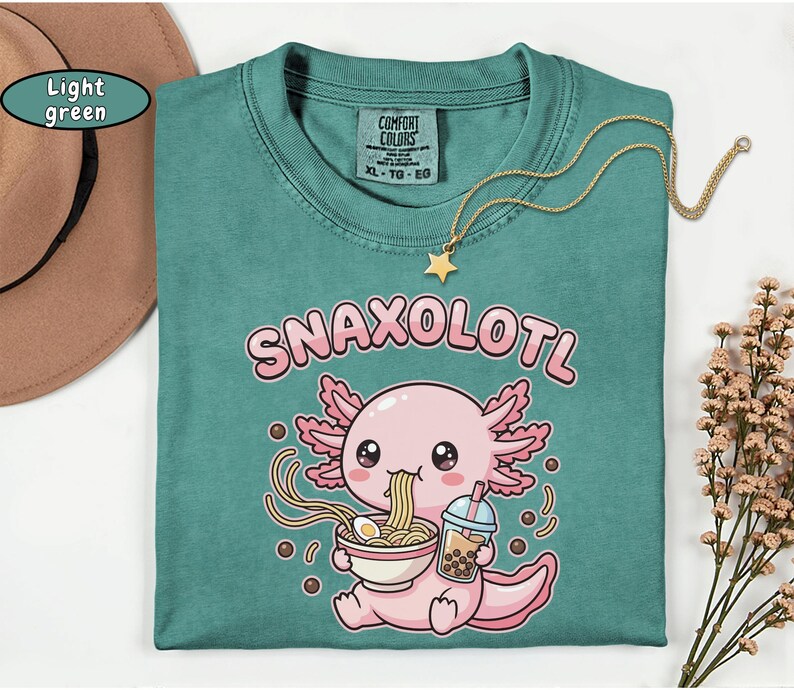 以下が含まれることがあります： 「Snaxolotl」の文字と、ラーメンを食べ、ボバを飲んでいる漫画のウーパールーパーが描かれた、ライトグリーンのComfort Colors Tシャツ。 金色の星のネックレスがシャツの上にかけられています。 茶色の帽子が左上にあります。