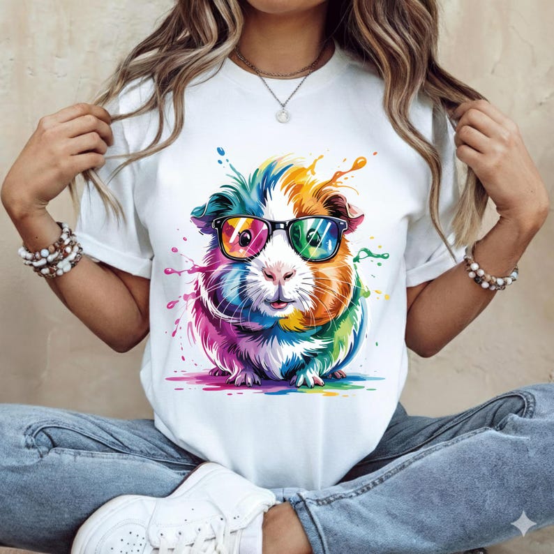 May include: White t-shirt featuring a colorful guinea pig wearing black-framed glasses. The guinea pig is depicted with vibrant hues of blue, orange, yellow, pink, and green, with paint splatter effects. The design is centered on the shirt.