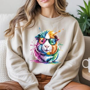 May include: Beige sweatshirt featuring a colorful guinea pig wearing glasses. The guinea pig is depicted with vibrant rainbow hues and watercolor paint splatters. The design is centered on the front of the sweatshirt.