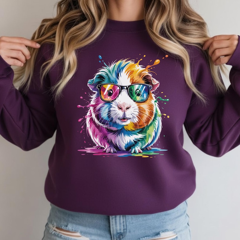 May include: A purple sweatshirt featuring a colorful guinea pig wearing sunglasses. The guinea pig is depicted with vibrant rainbow colors and paint splatter effects. The design is centered on the front of the sweatshirt.