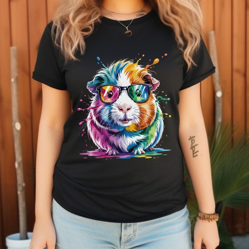 May include: Black t-shirt featuring a colorful guinea pig wearing glasses. The guinea pig is depicted with rainbow-colored fur and paint splatter effects, creating a vibrant and playful design.