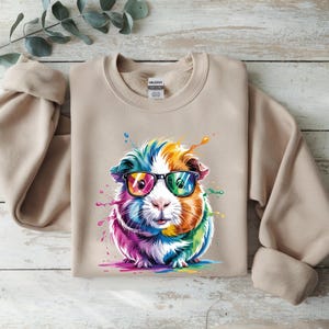 May include: A tan crewneck sweatshirt featuring a colorful graphic of a guinea pig wearing glasses. The guinea pig is depicted with rainbow-colored fur and paint splatters. The sweatshirt is made of a soft material.
