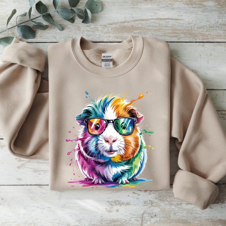 May include: A tan crewneck sweatshirt featuring a colorful graphic of a guinea pig wearing glasses. The guinea pig is depicted with rainbow-colored fur and paint splatters. The sweatshirt is made of a soft material.