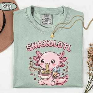 以下が含まれることがあります： 「Snaxolotl」の文字と、ラーメンを食べ、バブルティーを持っている漫画のウーパールーパーが描かれた、薄緑色のTシャツ。ウーパールーパーはピンク色で、ピンク色のフリルが付いています。星のペンダントが付いたゴールドのネックレスがTシャツの上にあります。