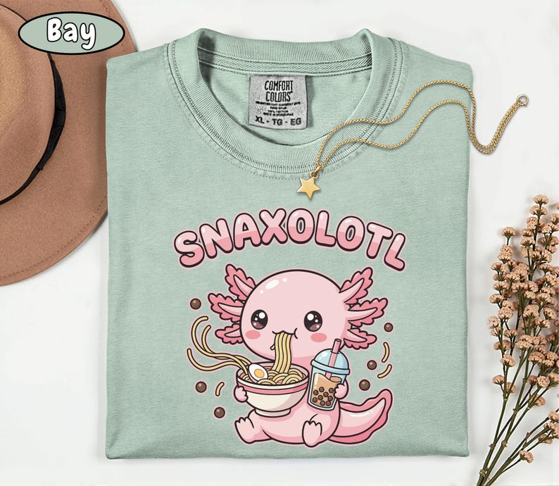 以下が含まれることがあります： 「Snaxolotl」の文字と、ラーメンを食べ、バブルティーを持っている漫画のウーパールーパーが描かれた、薄緑色のTシャツ。ウーパールーパーはピンク色で、ピンク色のフリルが付いています。星のペンダントが付いたゴールドのネックレスがTシャツの上にあります。