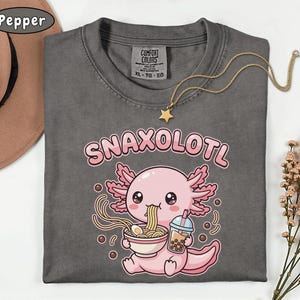 以下が含まれることがあります： 「SNAXOLOTL」の文字と、ラーメンを食べ、ボバドリンクを持っている漫画のウーパールーパーが描かれたダークグレーのTシャツ。星のペンダントが付いたゴールドのネックレスがシャツの上にかけられています。