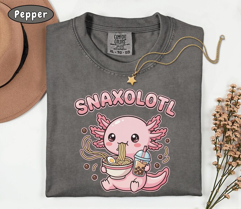 以下が含まれることがあります： 「SNAXOLOTL」の文字と、ラーメンを食べ、ボバドリンクを持っている漫画のウーパールーパーが描かれたダークグレーのTシャツ。星のペンダントが付いたゴールドのネックレスがシャツの上にかけられています。