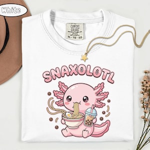 以下が含まれることがあります： 白いTシャツ。ピンク色のウーパールーパーがラーメンを食べ、ボバドリンクを持っているイラスト。テキスト「Snaxolotl」はウーパールーパーの上にあります。金の星のペンダントネックレスがTシャツの上にあります。