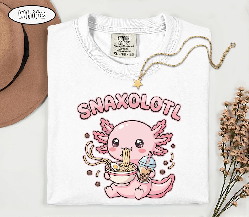 以下が含まれることがあります： 白いTシャツ。ピンク色のウーパールーパーがラーメンを食べ、ボバドリンクを持っているイラスト。テキスト「Snaxolotl」はウーパールーパーの上にあります。金の星のペンダントネックレスがTシャツの上にあります。