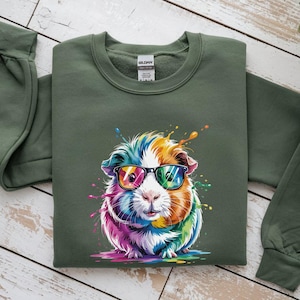 May include: An olive green sweatshirt featuring a colorful guinea pig wearing sunglasses. The guinea pig has a rainbow-colored coat with splashes of paint around it. The sweatshirt is made of a soft material.