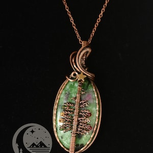 以下が含まれることがあります： 緑と赤の石を使った銅線ペンダントネックレス。ワイヤーで包まれたクリスマスツリーのデザインが特徴です。ペンダントは銅のチェーンから吊り下げられています。ネックレスは黒い背景にあります。