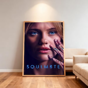 Peut inclure: Une affiche encadrée présentant le portrait en gros plan d'une personne aux yeux bleu vif et aux cheveux blonds. Une main, montrant des détails métalliques semblables à des circuits et des marques rouges, est tenue près du visage. Le texte SOULM8TE apparaît en lettres bleues lumineuses.