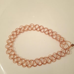 May include: A brown, stretchy choker necklace. The necklace is made of small, interconnected loops, creating a delicate, open design. The choker is laying on a white surface.