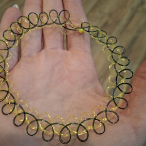 May include: A choker necklace with a spiral design. The necklace features alternating sections of black and yellow-gold colored plastic. The necklace is held in a person's hand.