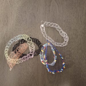 Puede incluir: Una colección de collares tipo gargantilla de tatuaje elásticos y una pulsera con cuentas. Las gargantillas son de varios colores, incluyendo transparente, negro y tonos pastel. La pulsera con cuentas presenta hilo azul y cuentas coloridas.