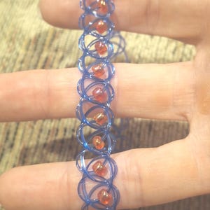 Puede incluir: Una pulsera azul con un diseño tejido, con pequeñas cuentas naranjas. La pulsera está hecha de un material flexible y está diseñada para ser usada en la muñeca. Las cuentas están espaciadas uniformemente.