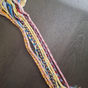 May include: A collection of colorful friendship bracelets. The bracelets are made of braided embroidery floss in various colors, including pink, yellow, blue, and red. The bracelets are arranged on a dark gray surface.