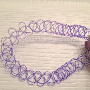 May include: A purple and pink choker necklace with a tattoo-style design. The necklace is made of a clear, stretchy material and features a repeating pattern of interlocking circles. The choker is shown against a white background.