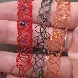 May include: Three wire-wrapped bracelets in red, black, and copper colors. Each bracelet features a unique design with small beads in various colors, including blue, green, and orange. The bracelets are displayed on a neutral background.