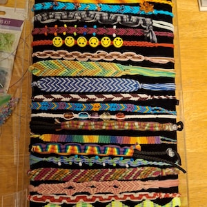 Puede incluir: Una colección de pulseras de la amistad coloridas exhibidas sobre una superficie de terciopelo negro. Las pulseras presentan varios patrones tejidos, cuentas y dijes de caritas sonrientes. La paleta de colores incluye azules, verdes, amarillos y rojos.