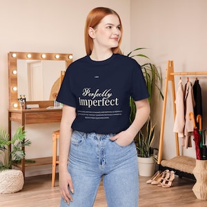 May include: Navy blue t-shirt with the text "Perfectly Imperfect" in a stylish font. The shirt also includes the text "I AM" at the top and a quote below the main text. The model is wearing blue jeans.