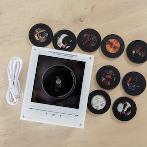 May include: A white square music player frame with a central circular opening, a white USB charging cable, and eight circular black discs featuring various album cover art. The frame displays the text THIS IS THE SHINING STAR IN MY ORDINARY LIFE.