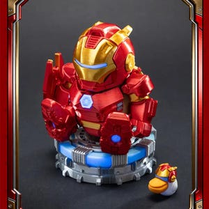 May include: A red and gold Iron Man figure with a blue light base and a small duck figure. The Iron Man figure has a detailed helmet and armour. The duck figure is wearing an Iron Man helmet. The figures are set against a dark background.