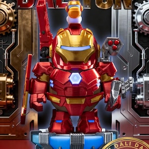 May include: A red and gold Iron Man-themed figurine with the word "DALTUN" in the background. The figurine has a red and gold suit, a blue light in the chest, and is holding a red weapon. The figurine is on a blue and grey platform.