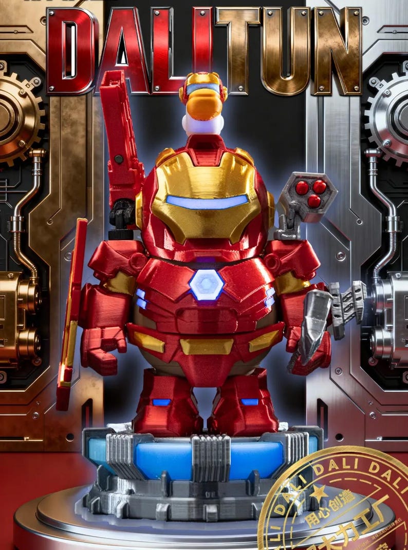May include: A red and gold Iron Man-themed figurine with the word "DALTUN" in the background. The figurine has a red and gold suit, a blue light in the chest, and is holding a red weapon. The figurine is on a blue and grey platform.