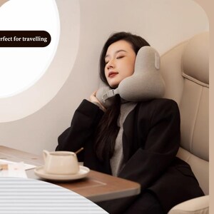May include: A gray travel neck pillow is around a person's neck. The person is sitting in a plane seat, with a cup of coffee on a tray table. The text "Perfect for travelling" is in the upper left corner.