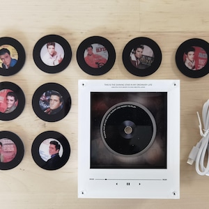May include: A white square music player frame with a central circular opening, eight black circular coasters featuring Elvis Presley portraits, and a white USB power cable. The frame displays the text "THIS IS THE SHINING STAR IN MY ORDINARY LIFE" and "PLEASE PUT IN THE MUSIC ALBUM YOU WANT TO PLAY" with playback icons.